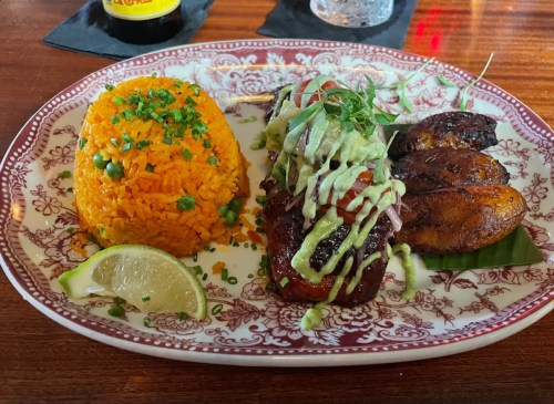 A plate featuring orange rice, grilled plantains, and a meat dish topped with green sauce and garnished with herbs.