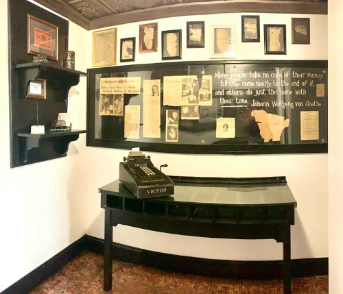 A vintage typewriter on a desk in a room with framed photos and newspaper clippings on the walls.