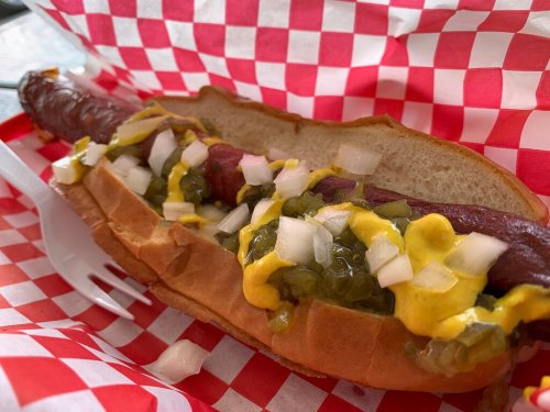 A hot dog in a bun topped with mustard, chopped onions, and pickles, served in a red and white checkered paper.