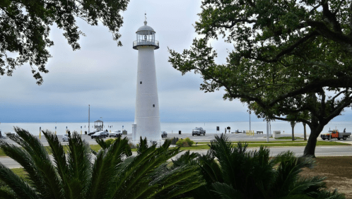 A tall white lighthouse stands by the shore, surrounded by trees and palm plants, under a cloudy sky.