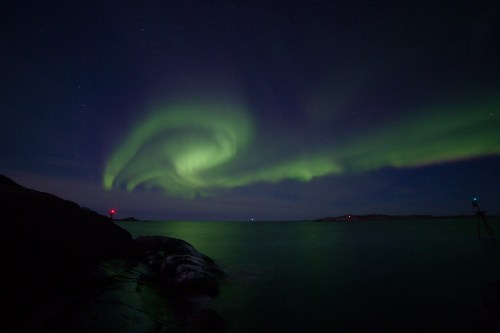 A mesmerizing display of the Northern Lights swirls over a calm sea, with faint lights on the horizon and rocky shore.