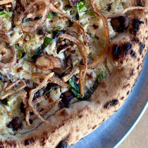A close-up of a pizza topped with herbs, crispy onions, and chunks of meat on a charred crust.