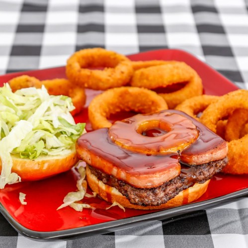 A plate with a burger topped with a donut and barbecue sauce, served with onion rings and a side of lettuce.