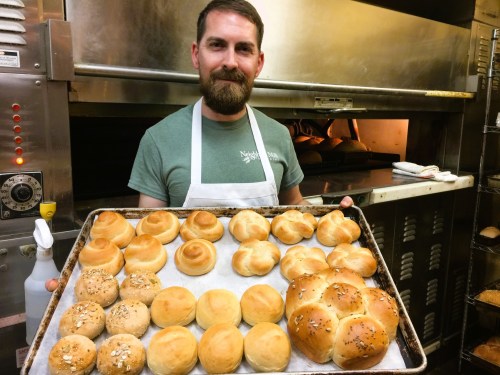 A baker holds a tray of freshly baked rolls in a kitchen, with an oven in the background.