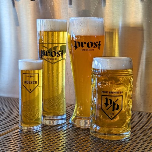 Four glasses of beer in various sizes, all featuring the "Prost Brewing Co" logo, on a metal countertop.