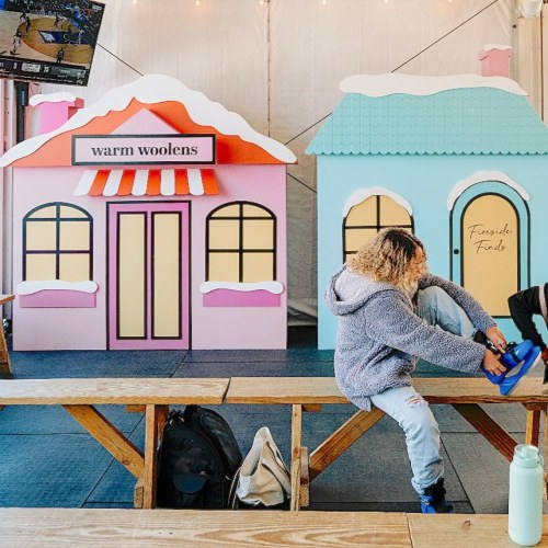 A person sits on a bench, putting on shoes near colorful, whimsical storefronts in a festive setting.