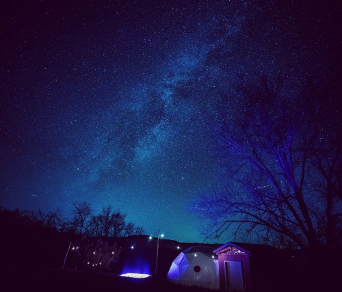 A starry night sky with the Milky Way, featuring a dome structure and a small building illuminated by soft lights.