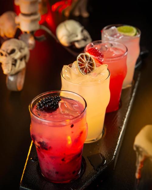 Four colorful cocktails with ice, garnished with fruit, on a dark table decorated with skulls.