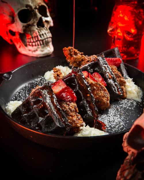A dark plate with waffles topped with strawberries and syrup, surrounded by skull decorations and a moody red background.