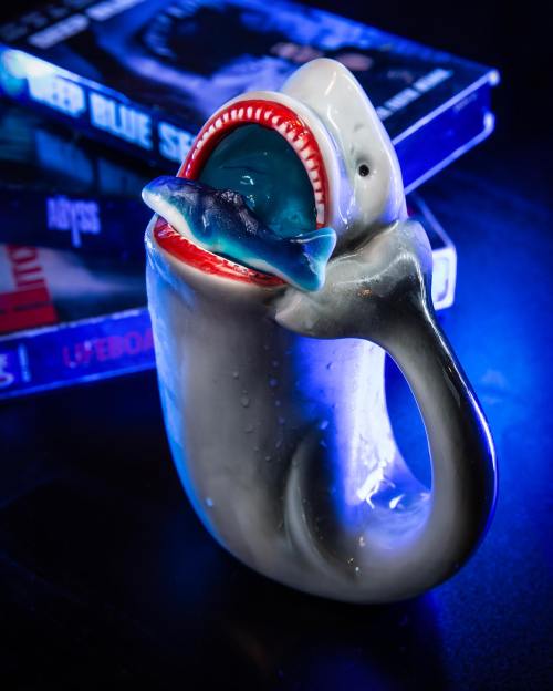 A shark-shaped mug with an open mouth, holding a small fish, set against a backdrop of blue-lit movie covers.