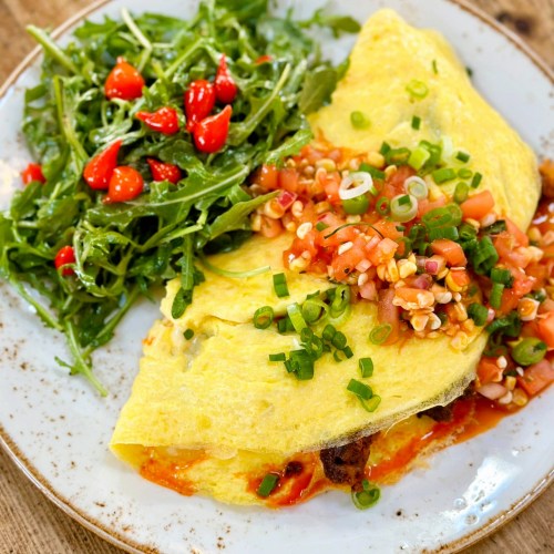 A fluffy omelette topped with salsa and green onions, served with a side of arugula and cherry tomatoes.