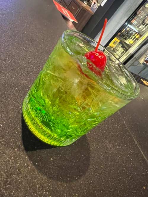 A vibrant green cocktail in a textured glass, topped with a cherry, sits on a dark countertop.