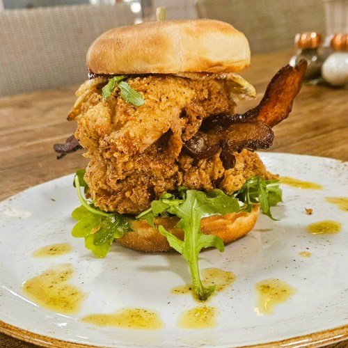 A towering fried chicken sandwich with bacon, lettuce, and a soft bun, drizzled with sauce on a plate.