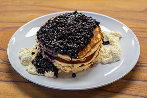 Stack of pancakes topped with a generous layer of blueberry sauce, served with a side of creamy ricotta on a white plate.