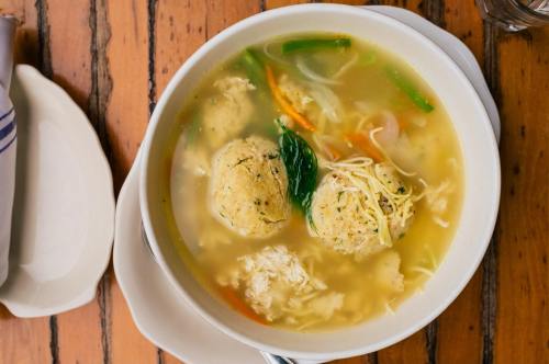 A bowl of soup with dumplings, vegetables, and herbs, served on a wooden table.