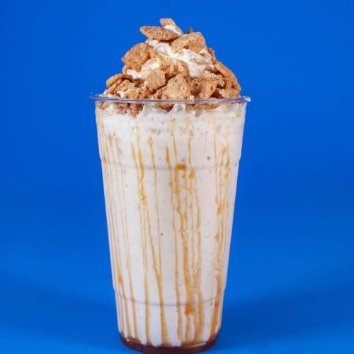 A tall cup filled with a creamy milkshake topped with cereal and caramel drizzle against a blue background.
