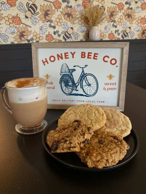 A cozy café scene featuring a latte, a plate of cookies, and a sign for Honey Bee Co. with a bicycle graphic.