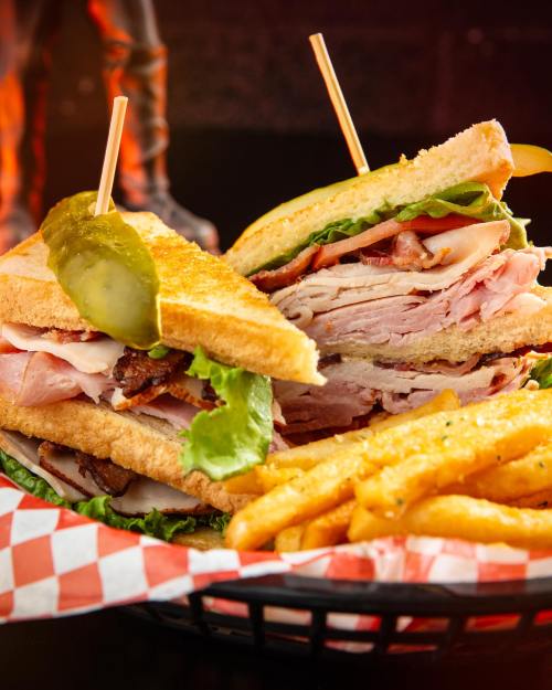 A stacked sandwich with layers of meats, lettuce, and pickles, served with golden fries in a checkered basket.