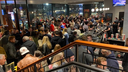 A crowded bar scene with many people socializing, drinks on tables, and a lively atmosphere.