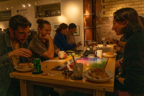 A group of people at a table playing a board game, enjoying food and drinks in a cozy, well-lit space.