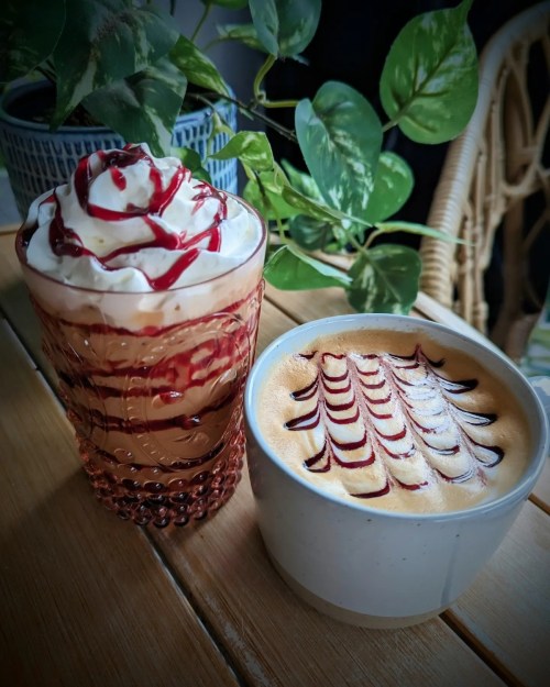A glass of layered dessert topped with whipped cream and syrup, alongside a cup of coffee with artistic foam design.
