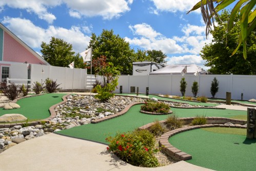 A sunny mini-golf course with green turf, rocks, and landscaping, surrounded by trees and a blue sky with clouds.