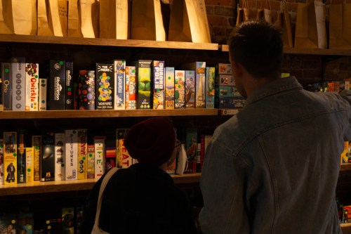 Two people browse a shelf filled with colorful board games in a cozy store.