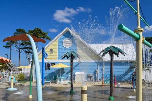 A colorful splash pad with palm tree features, water sprays, and a blue building under a clear sky.
