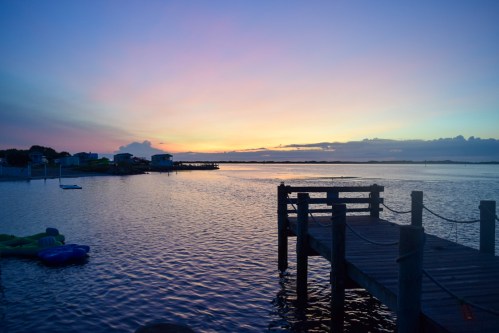 A serene sunset over a calm waterway, with a wooden dock and colorful sky reflecting on the water.