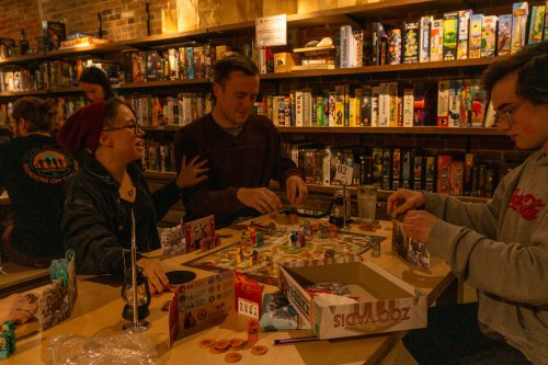 A group of friends plays a board game at a cozy game cafe, surrounded by shelves of board games.