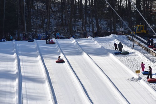 A snowy hill with people tubing down, surrounded by trees and a ski lift in the background.