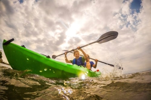 Two children paddle a bright green kayak through water under a cloudy sky, splashing as they row.
