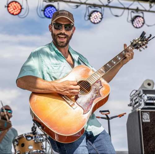 A musician in a light blue shirt plays an acoustic guitar on stage, smiling and enjoying the performance.