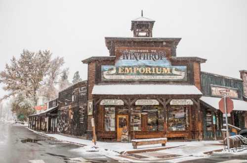 A snowy scene of the Winthrop Emporium, a rustic building with a sign, surrounded by winter scenery.