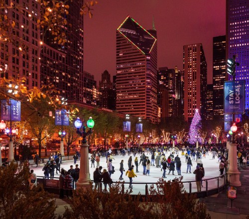 A bustling ice skating rink surrounded by city skyscrapers, illuminated by colorful lights and a festive tree at night.