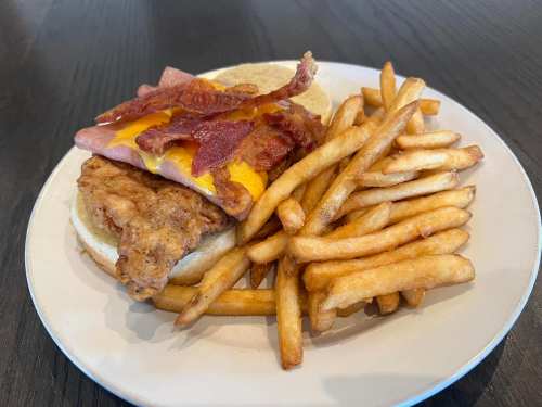 A plate with a sandwich featuring fried chicken, cheese, bacon, and a side of golden French fries.