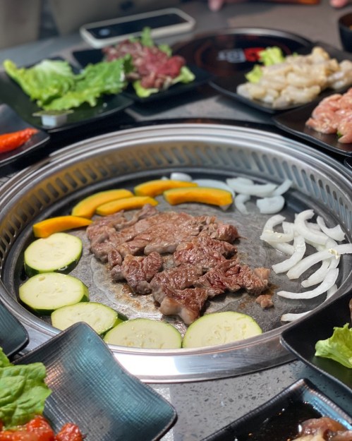 Grilled meat on a hot plate surrounded by fresh vegetables and various side dishes on a table.