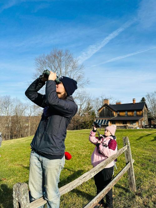 Two people outdoors, one using binoculars and the other taking photos, with a house and trees in the background.