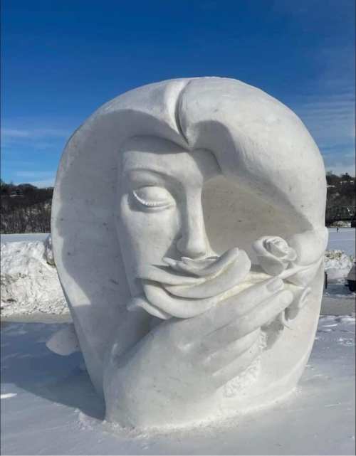 A large white snow sculpture of a woman's face gently holding a rose, set against a clear blue sky.