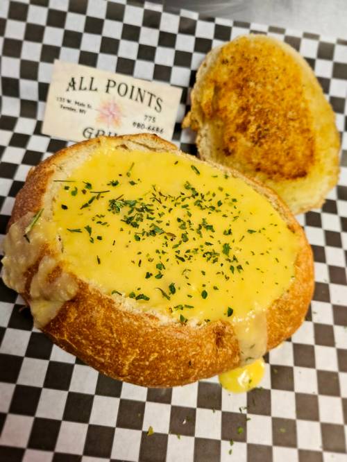 A bread bowl filled with creamy cheese sauce, garnished with herbs, next to a toasted bread top on a checkered paper.