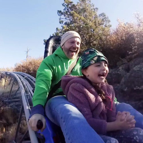 A man and a girl joyfully ride a roller coaster through a scenic outdoor landscape, both smiling and excited.