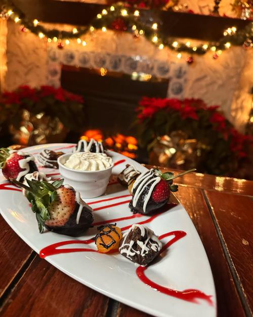 A festive plate of chocolate-covered strawberries and a small dessert, set against a cozy, decorated fireplace backdrop.