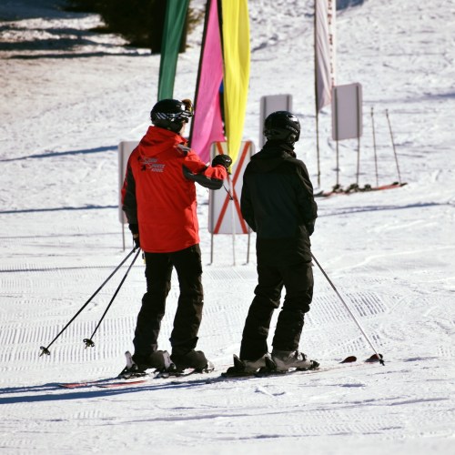 Two skiers on a snowy slope, one wearing a red jacket and the other in black, preparing to ski. Colorful flags in the background.