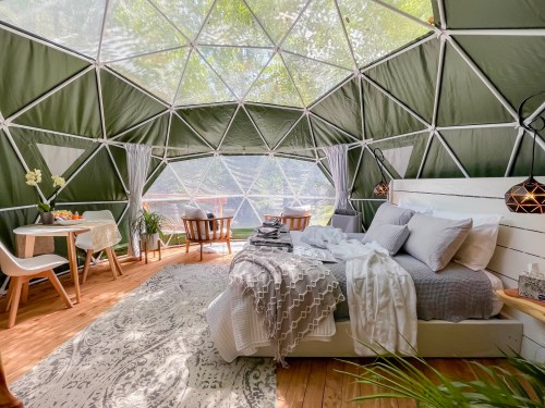 Cozy interior of a geodesic dome with a bed, dining area, and large windows overlooking nature.