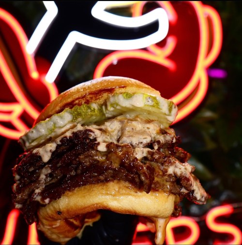 A close-up of a juicy burger with pickles and sauce, set against a vibrant neon background.