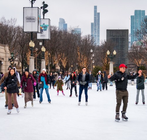 A bustling ice skating rink with people skating, surrounded by trees and city skyscrapers in the background.