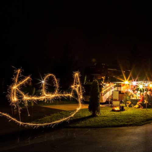 A nighttime scene with people gathered around a campfire, illuminated by sparklers, with a camper in the background.