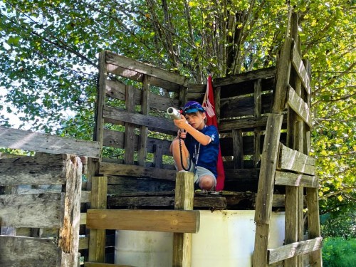 A child sits on a wooden fort, aiming a toy gun, surrounded by trees and a flag in the background.
