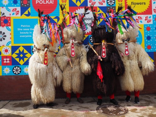 Four individuals in colorful, furry costumes with masks and decorative headdresses stand against a vibrant mural backdrop.