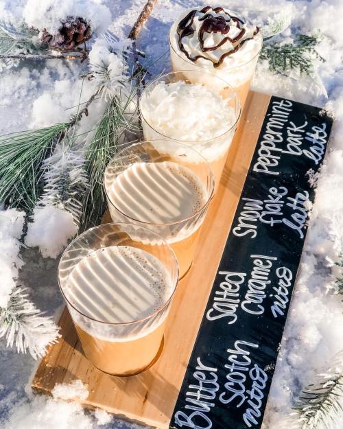A wooden tray with four cups of flavored lattes topped with whipped cream, set against a snowy backdrop.
